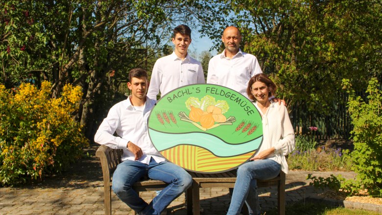 Vier Personen posieren mit einem Schild von 'Bachl's Feldgem&uuml;se' in einem Garten.