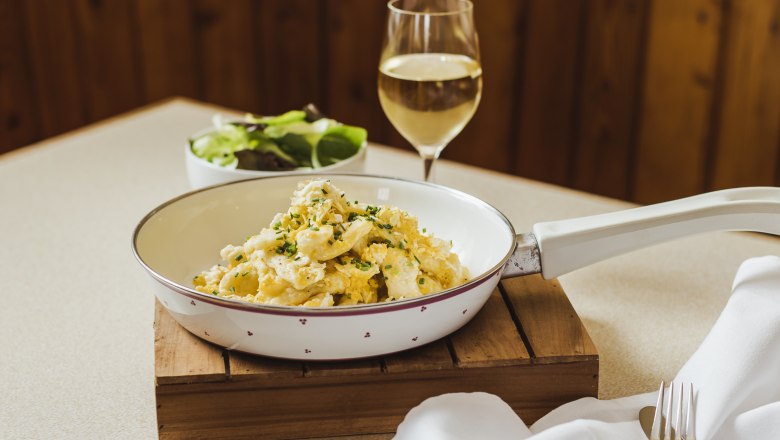 Eiernockerl in einer Pfanne mit einem Glas Wei&szlig;wein und einem Salat im Hintergrund.