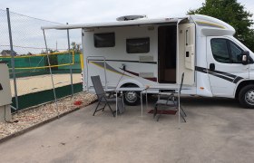 Wohnmobil mit ausgefahrener Markise und Campingm&ouml;beln auf einem Stellplatz neben einem Volleyballfeld.