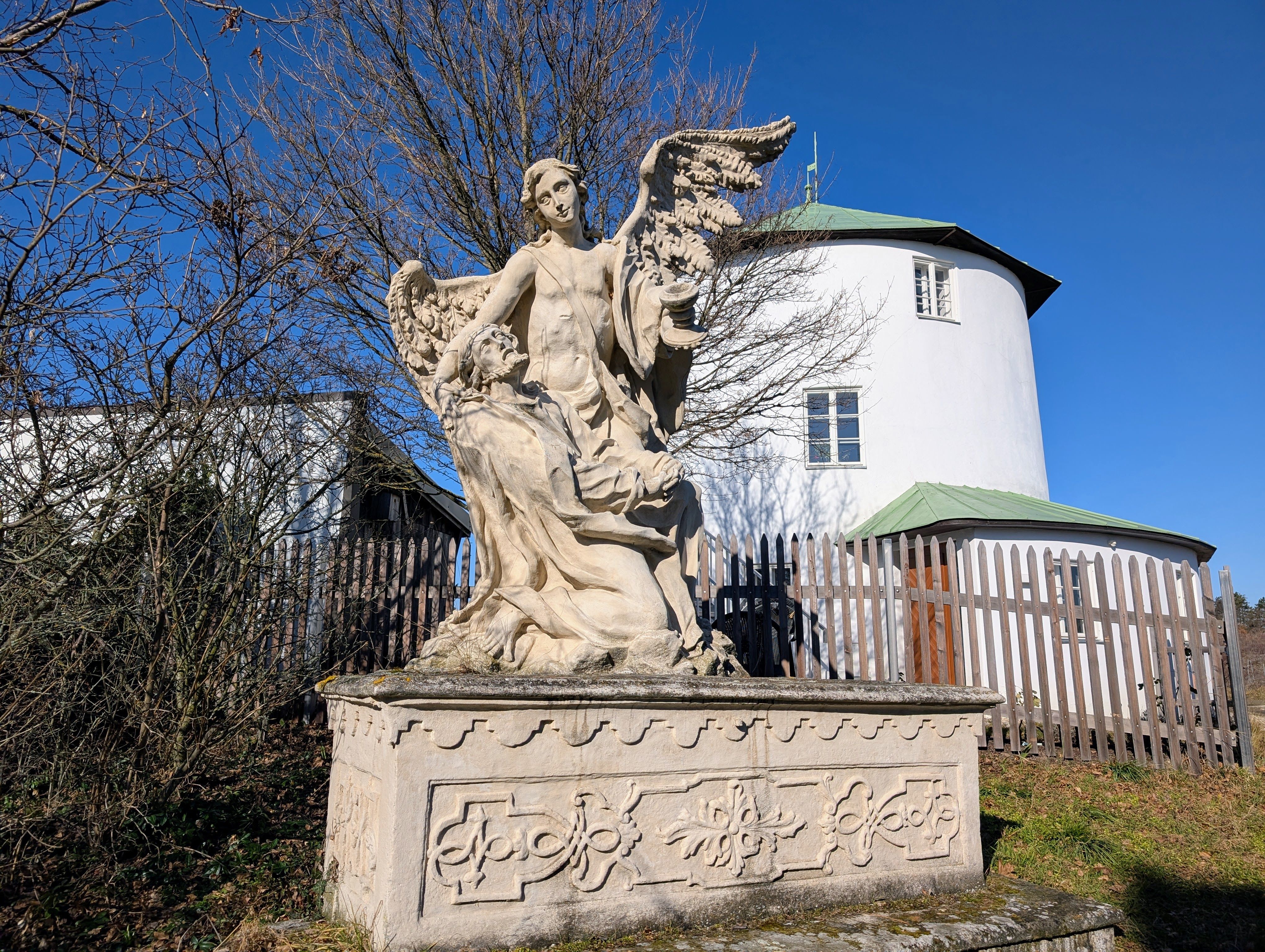 Steinstatue eines Engels mit Flügeln und einer knienden Figur vor einem runden, weißen Gebäude mit grünem Dach.