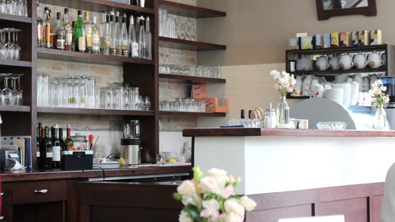 Innenansicht einer Bar mit Regalen voller Gläser und Flaschen, Blumen auf dem Tresen.