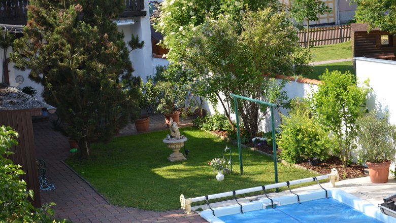 Ein gepflegter Garten mit Bäumen, Sträuchern, einer Statue und einem Pool. Im Hintergrund sind ein Haus und ein Zaun zu sehen.