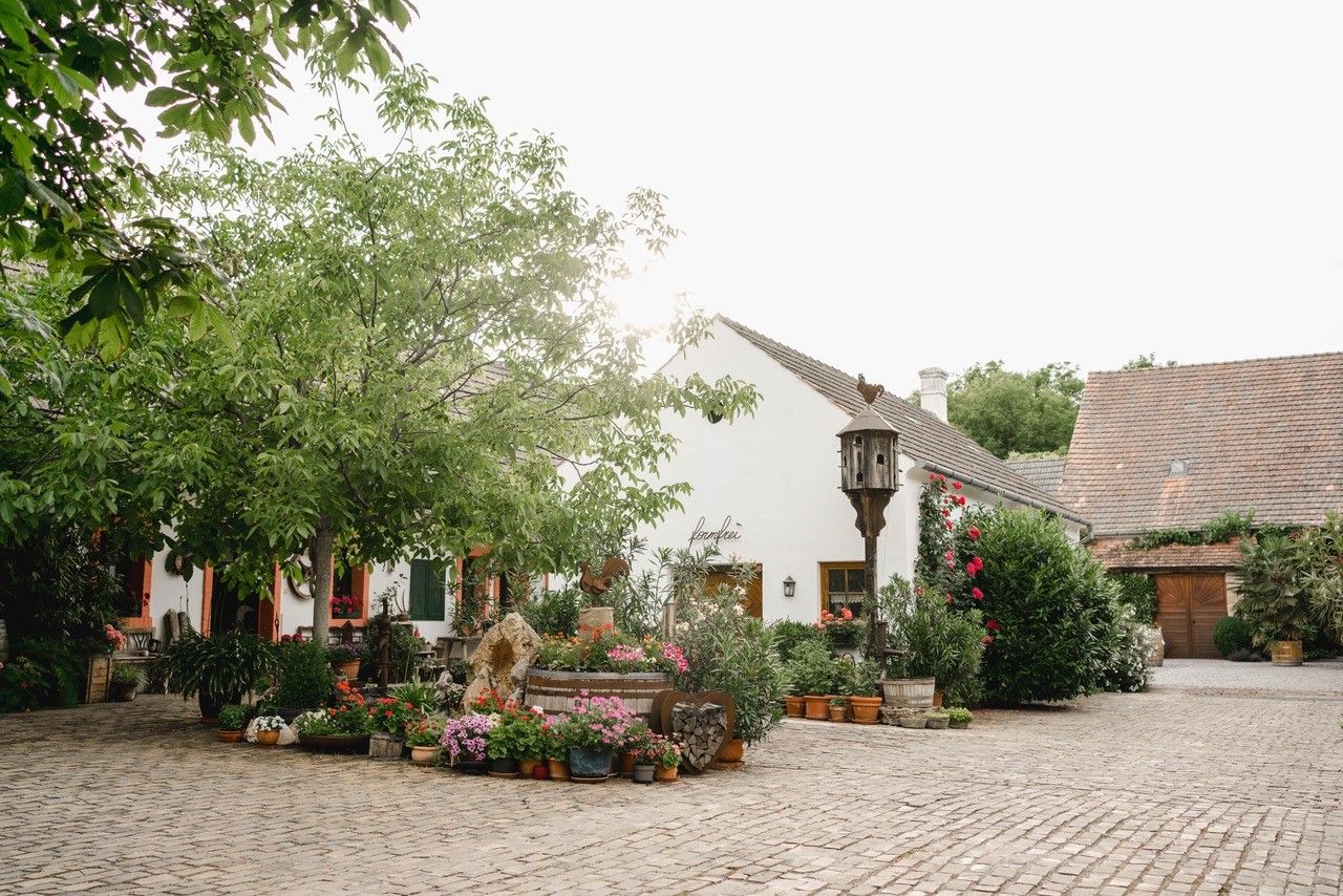 Ein idyllischer Hof mit Kopfsteinpflaster, umgeben von grünen Bäumen und bunten Blumen, vor einem weißen Gebäude mit Ziegeldach.