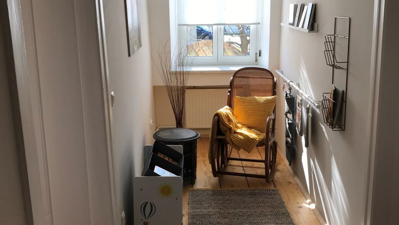 Ein schmaler Eingangsbereich mit Schaukelstuhl, gelbem Kissen, Teppich und Regalen an der Wand. Ein Fenster am Ende des Flurs.