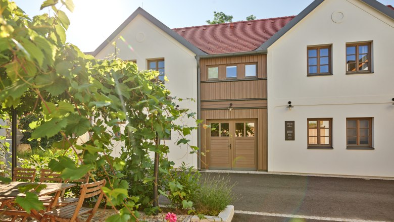 A modern, two-storey building with white walls and red roof tiles. In the foreground, a garden with a wooden table, chairs and flowering plants.
