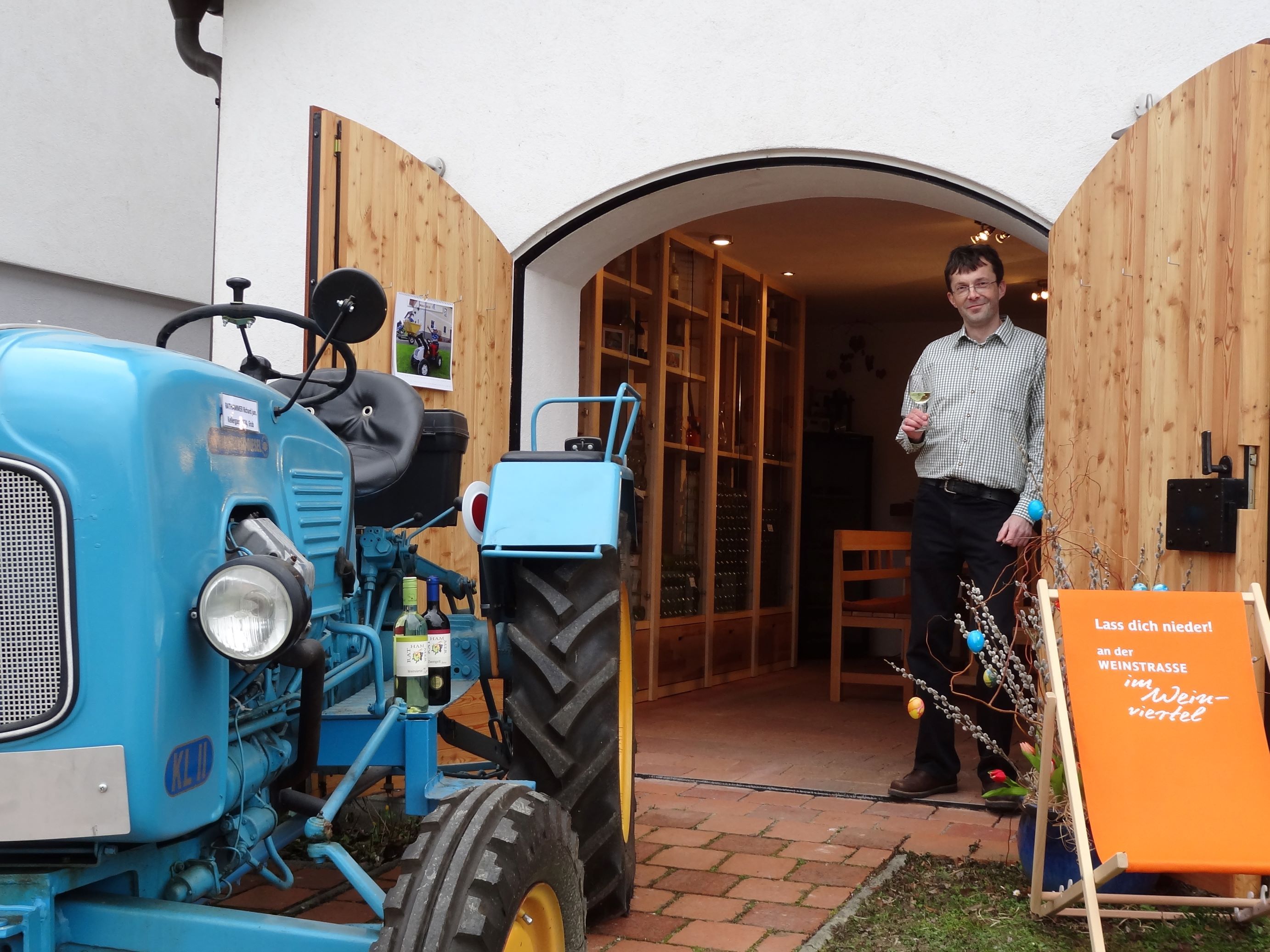 A man stands with a wine glass in his hand in front of a building with open wooden doors. A blue tractor is parked next to it.
