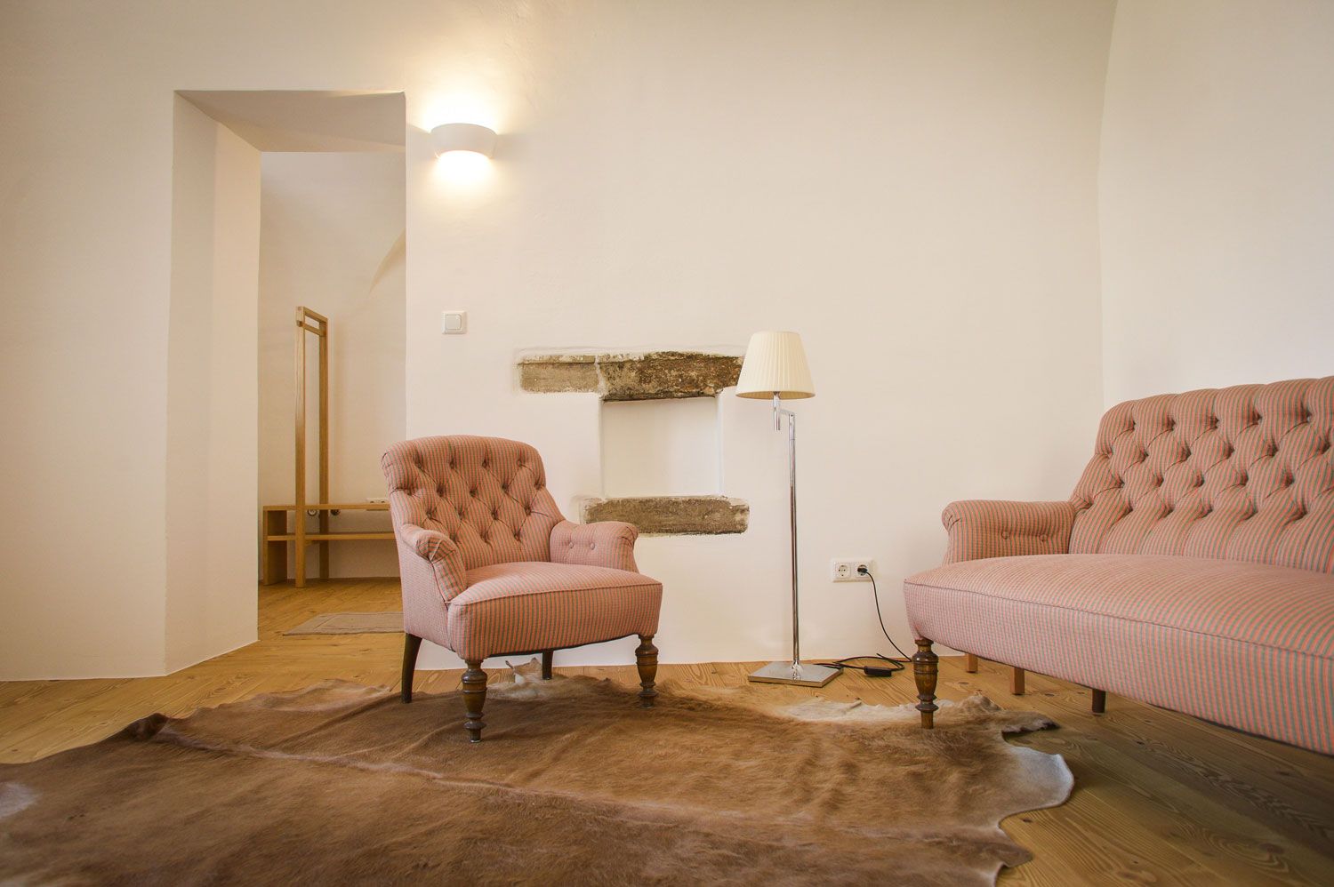 Ein gemütlicher Raum mit einem rosa gestreiften Sessel und Sofa, einer Stehlampe und einem Fellteppich auf Holzboden.