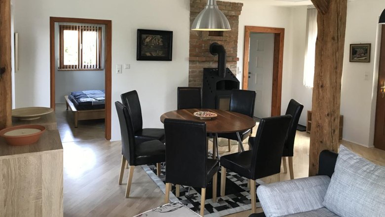 Essbereich mit rundem Holztisch, schwarzen Stühlen und Holzofen in einem Raum mit Holzbalken.