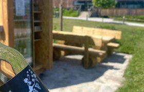 Eine Flasche mit der Aufschrift 'Spritzer rocks' vor einem Rastplatz mit Holzbänken und einem Kühlschrank im Freien.