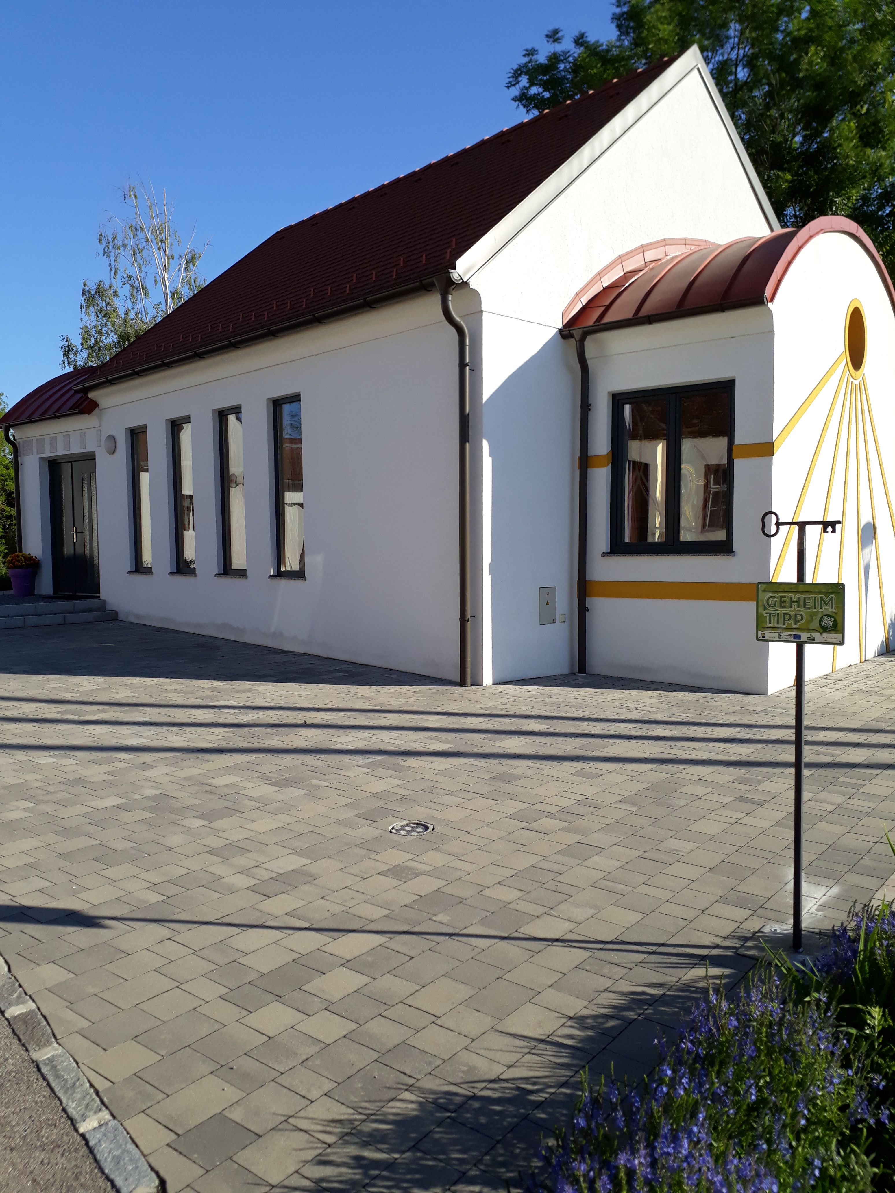 Weiße Kapelle mit rotem Dach und gelben Akzenten, umgeben von gepflastertem Boden und Pflanzen.