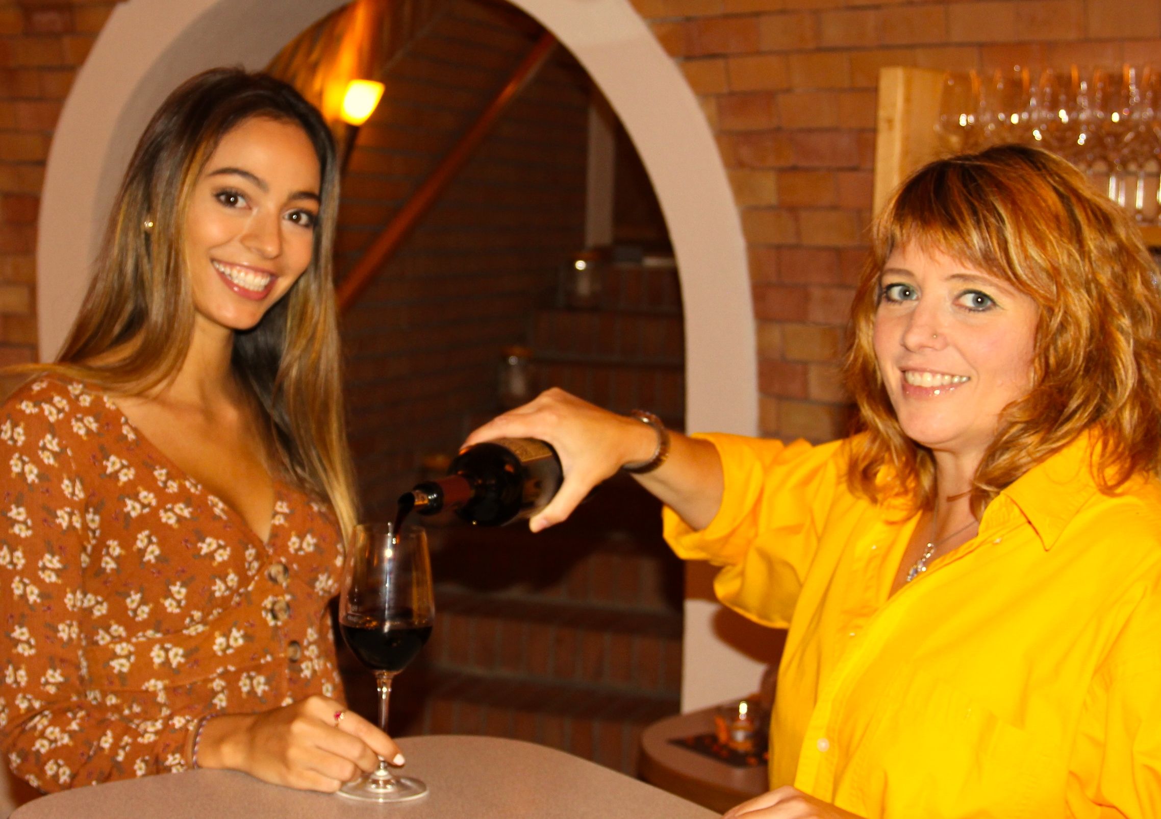Two women in a wine cellar, one pouring wine for the other.