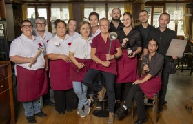 Ein Team von Restaurantmitarbeiterinnen und Mitarbeitern posiert in Arbeitskleidung mit Küchenutensilien in einem Speiseraum.