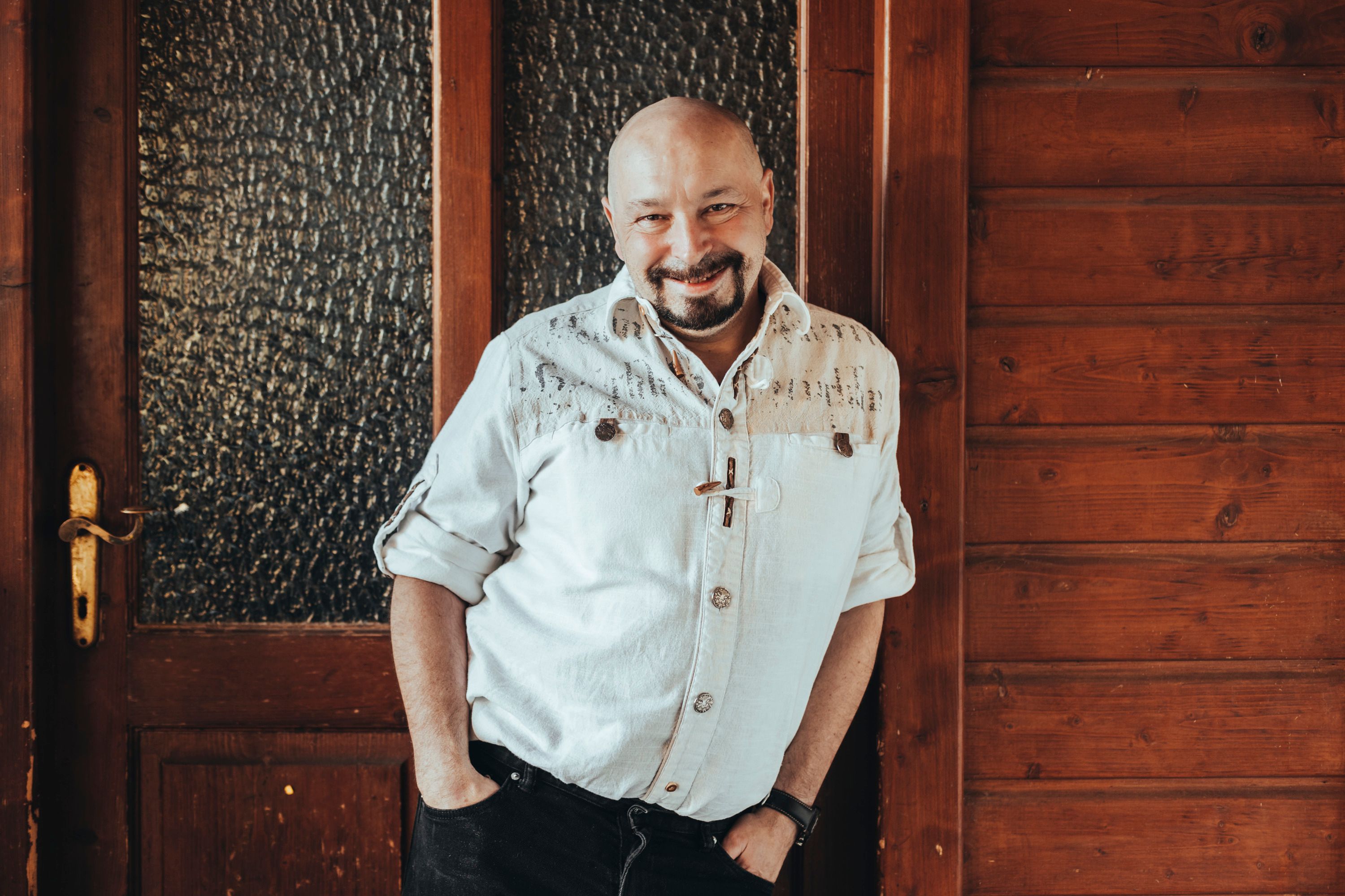 Ein Mann in einem weißen Hemd lehnt lächelnd an einer Holzwand.