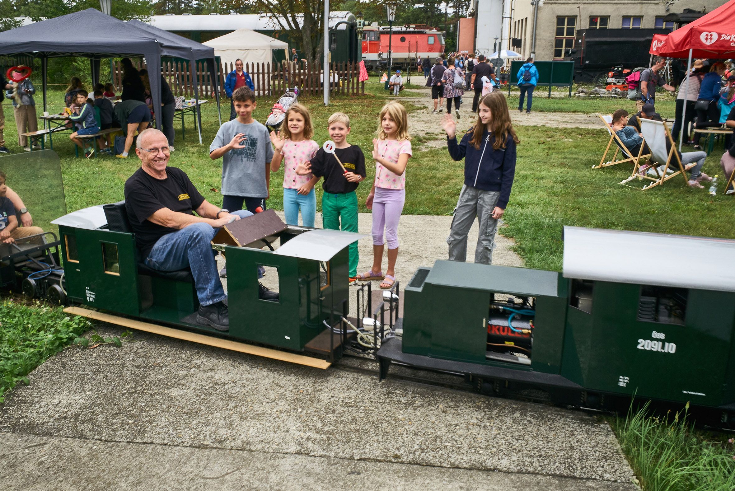 Ein Mann fährt eine Miniatur-Eisenbahn, während Kinder daneben stehen und winken. Im Hintergrund sind Zelte und weitere Besucher zu sehen.