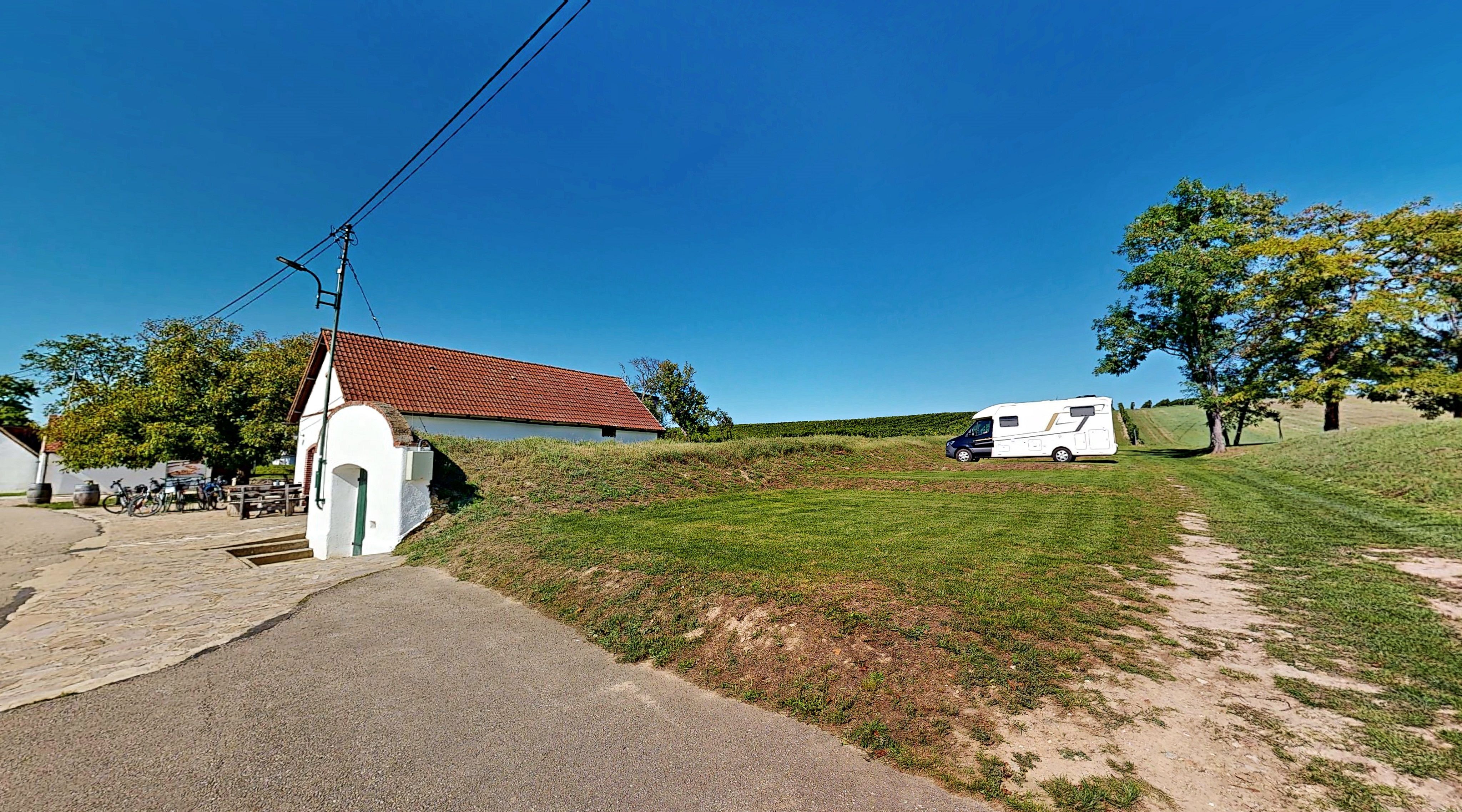 Ein Wohnmobil steht auf einem grünen Campingplatz neben einem kleinen weißen Gebäude mit rotem Dach. Im Hintergrund sind Bäume und blauer Himmel zu sehen.