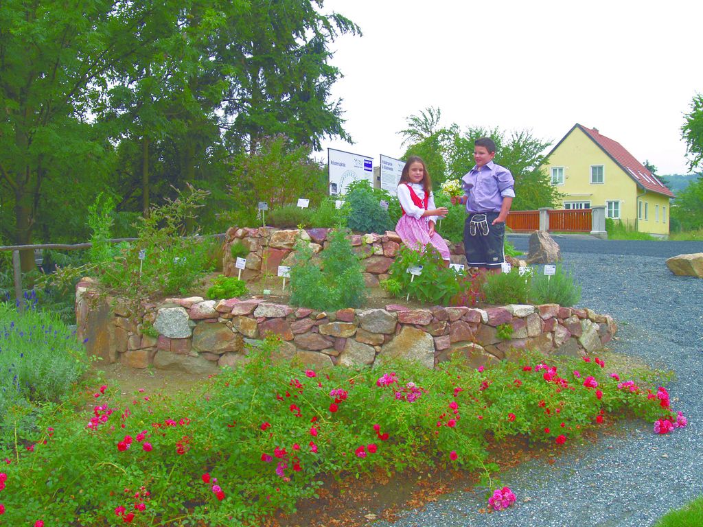 Zwei Kinder stehen in einer Kräuterspirale aus Steinen, umgeben von blühenden Pflanzen und einem gelben Haus im Hintergrund.