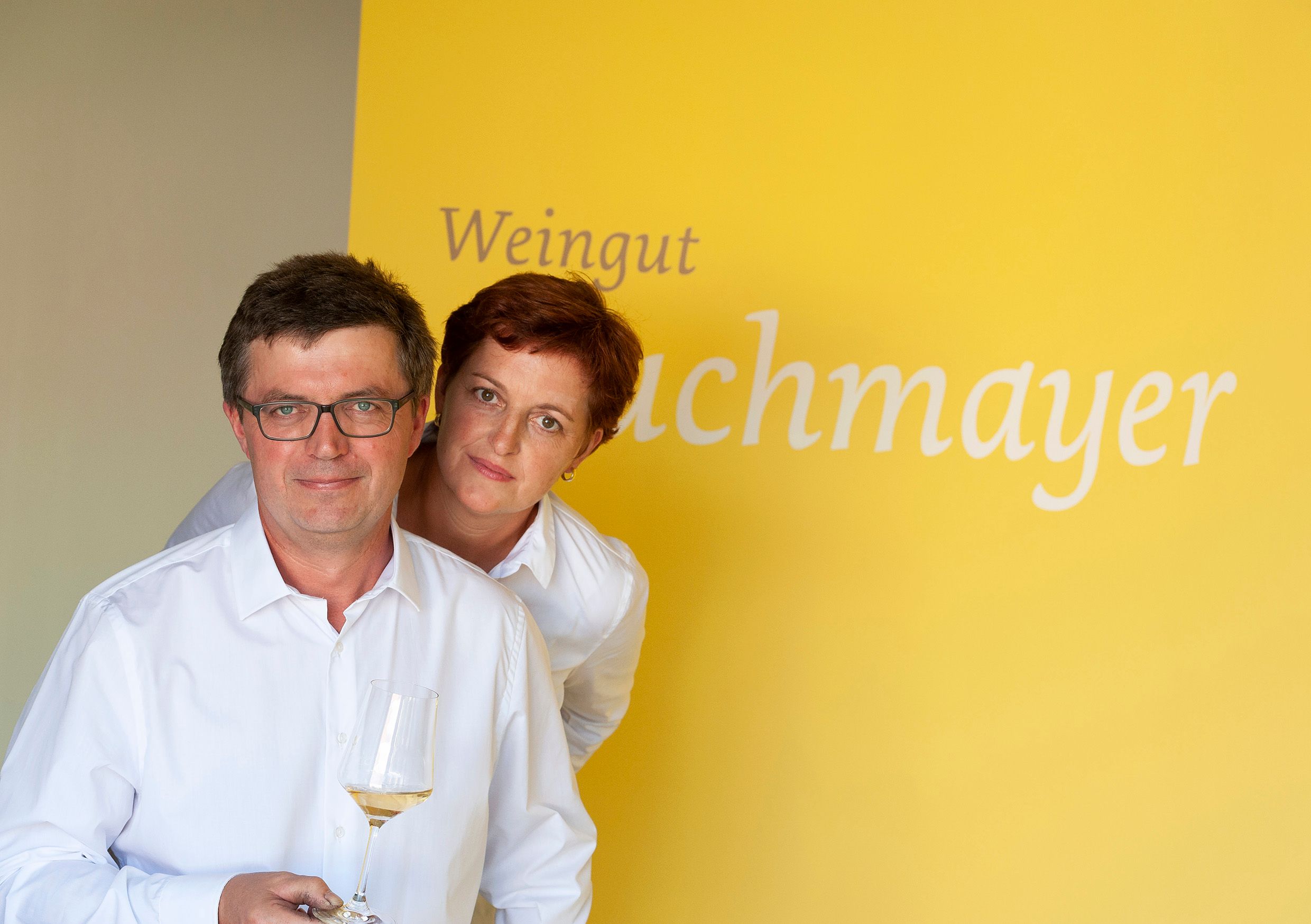 Dvě osoby v bílých košilích na žlutém pozadí s nápisem "Weingut Buchmayer". Jedna osoba drží v ruce sklenku na víno.