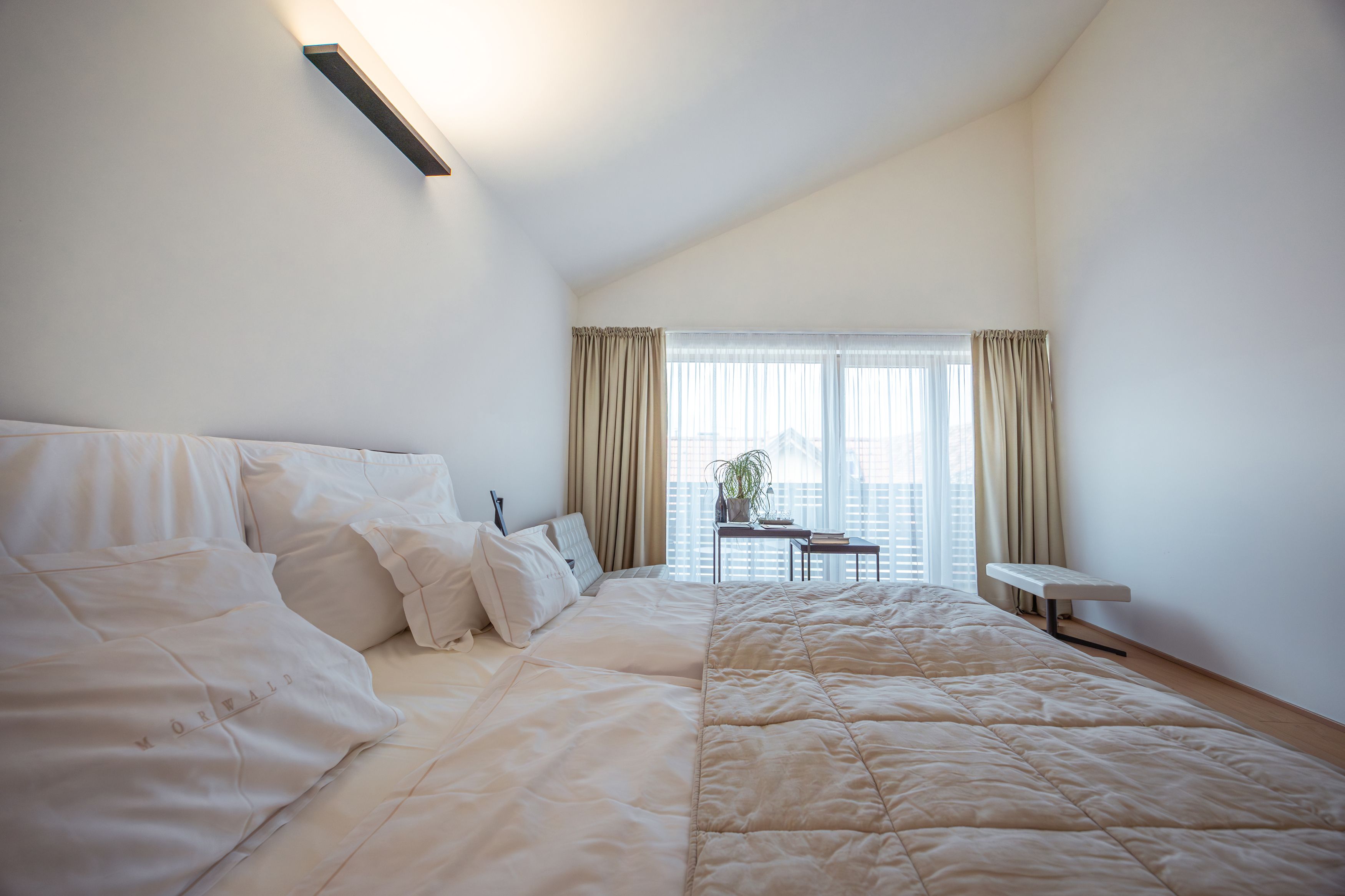 Modernes Hotelzimmer mit großem Bett, weißen Kissen und Tagesdecke, helle Vorhänge und Fenster im Hintergrund.