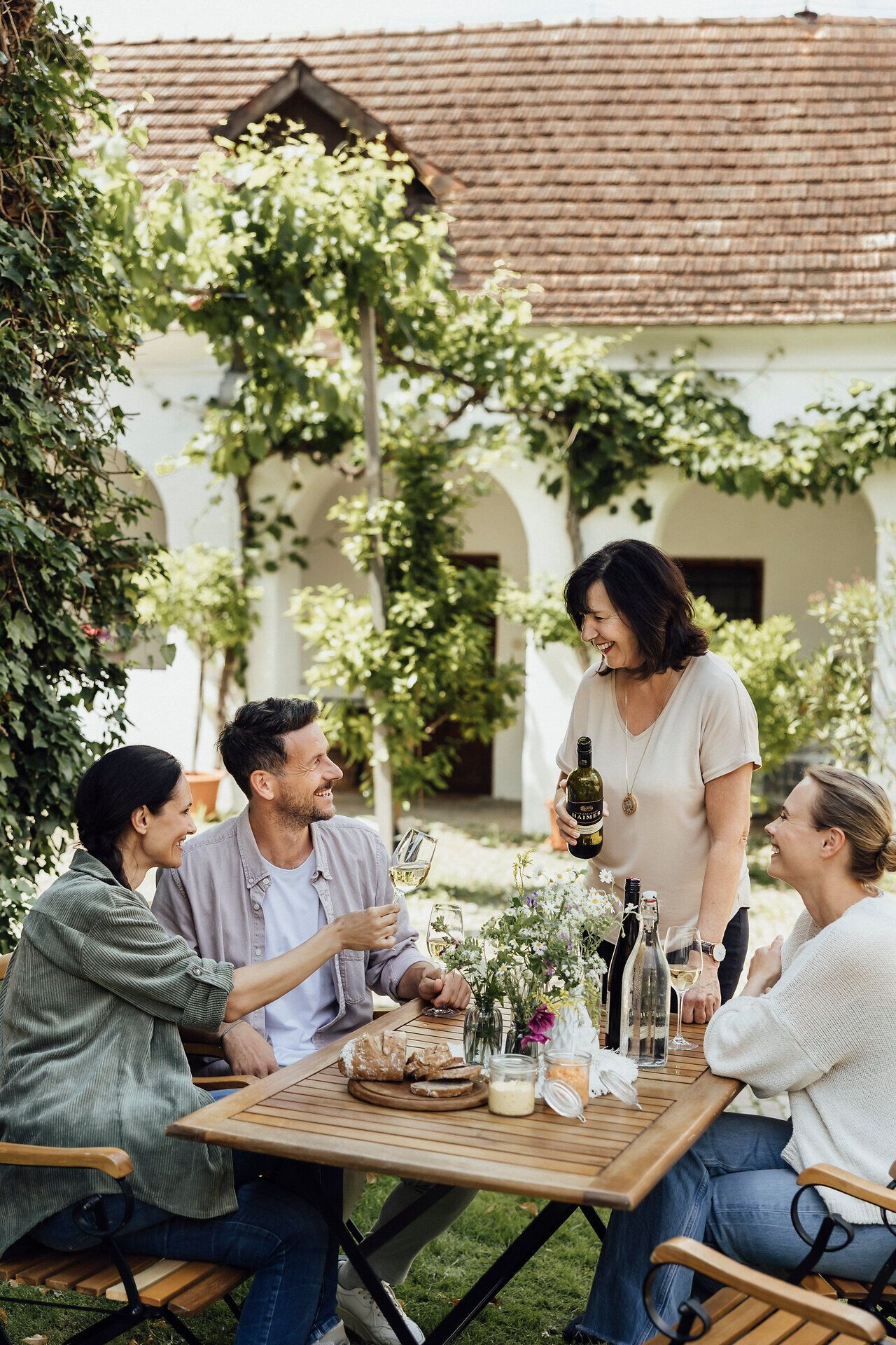 Inmitten einer malerischen Kulisse genießen Freunde eine gesellige Weinverkostung auf der Terrasse eines charmanten Winzerhofs. Umgeben von üppigem Grün und blühenden Pflanzen stoßen sie fröhlich mit Gläsern an, während eine köstliche Jause auf dem Tisch bereitsteht. Der Duft von frischem Wein und die entspannte Atmosphäre laden dazu ein, den Moment in vollen Zügen zu genießen.