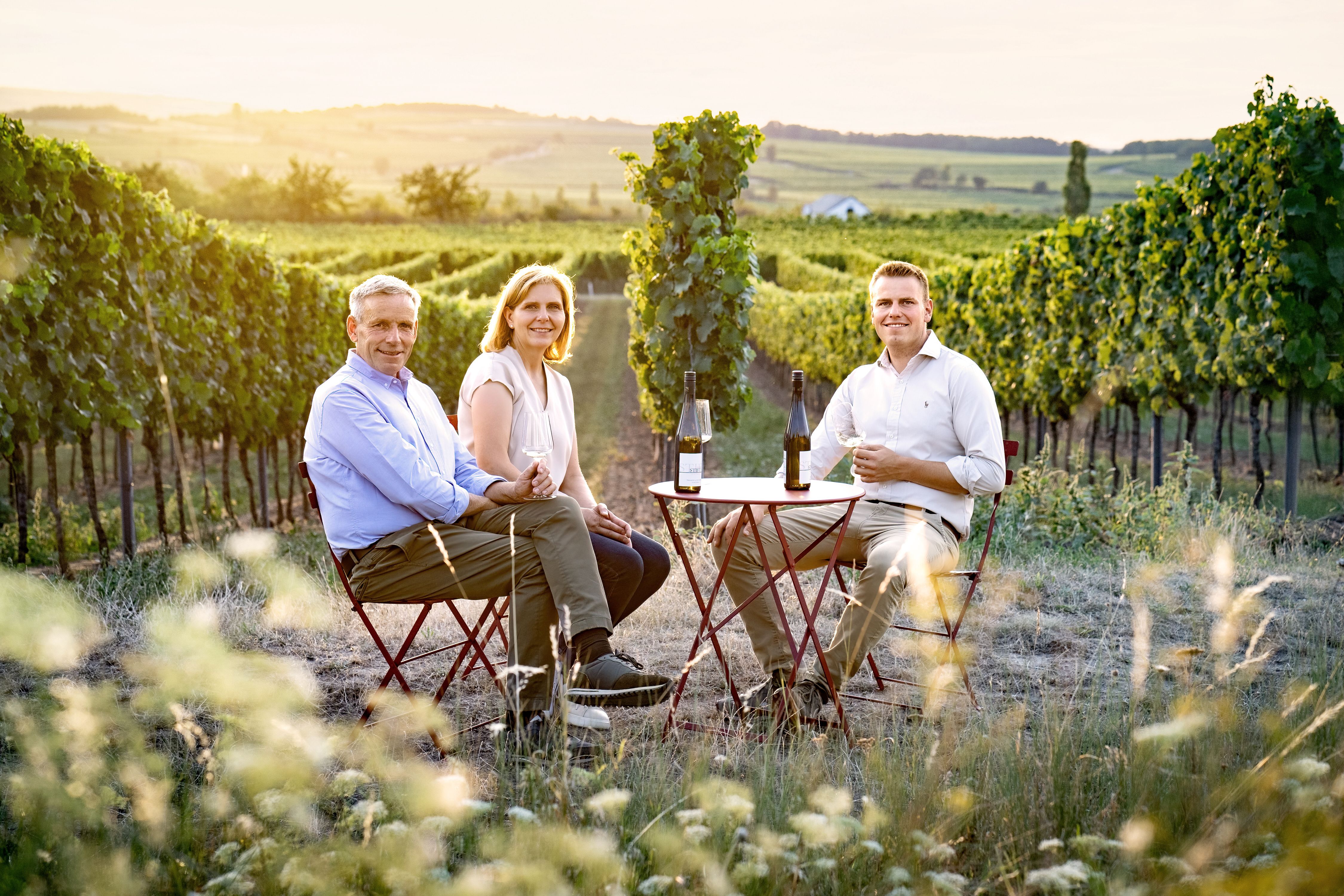 Eine Winzerfamilie - bestehend aus 3 Personen - sitzt an einem Tisch umgeben von Weingärten.