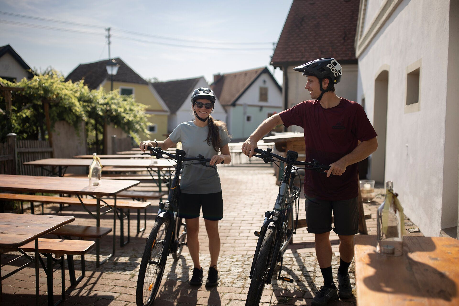 Zwei Radfahrer genießen die warme Sommerbrise in einer malerischen Kellergasse, umgeben von üppigem Grün und charmanten Weinhäusern. Die entspannte Atmosphäre lädt dazu ein, die Schönheit der Natur und die regionalen Köstlichkeiten zu entdecken.