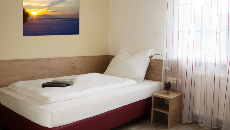 Ein schlichtes Einzelzimmer mit einem Bett, einem Nachttisch und einem Bild an der Wand. Ein Fenster mit Vorh&auml;ngen l&auml;sst Licht herein.