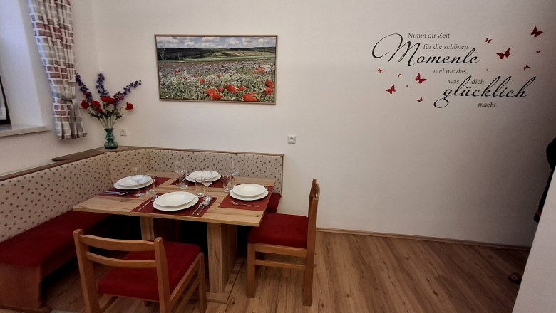 Essbereich mit Eckbank, Tisch und Stühlen, gedeckt mit Geschirr. An der Wand ein Bild von Mohnblumen und ein Zitat mit Schmetterlingen.