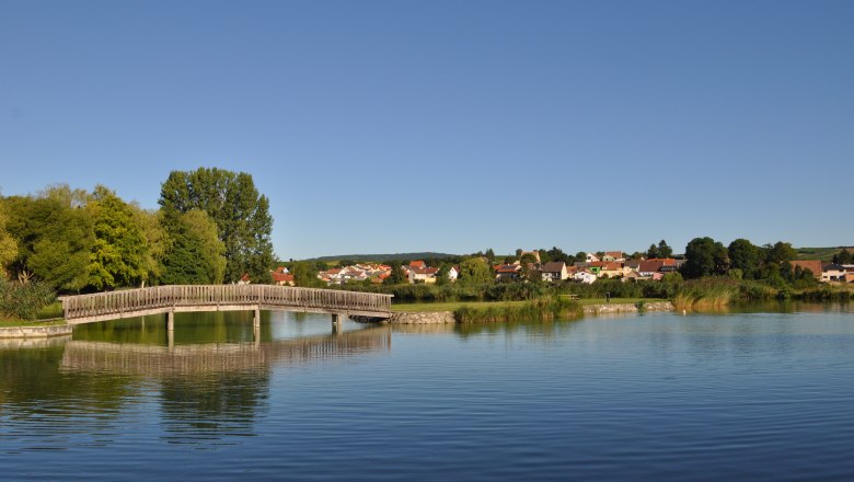 Holzbrücke über einen ruhigen Teich mit Dorf im Hintergrund.