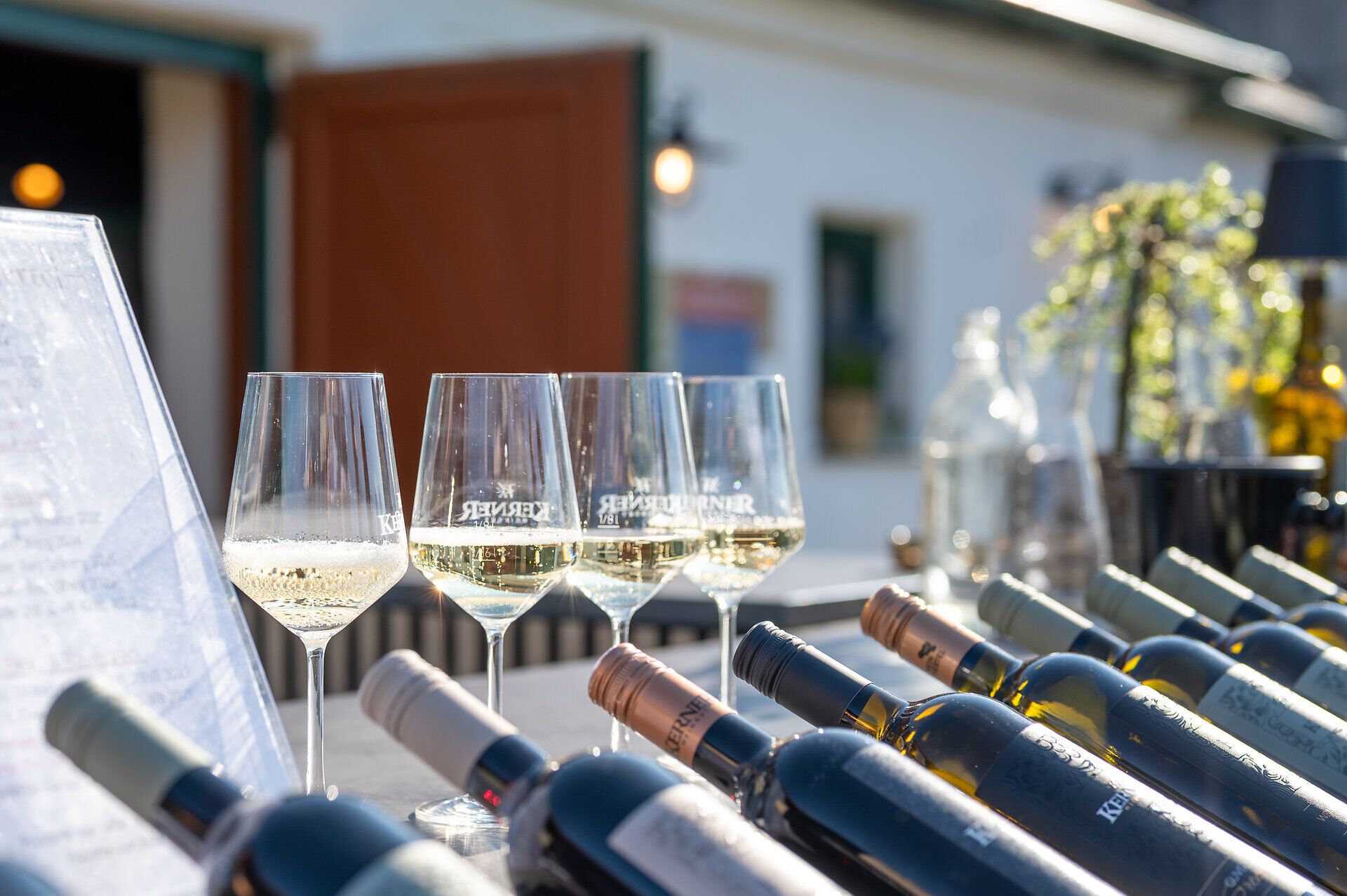 Vor einem Presshaus sind Weinflaschen und mit Weißwein gefüllte Weingläser positioniert.