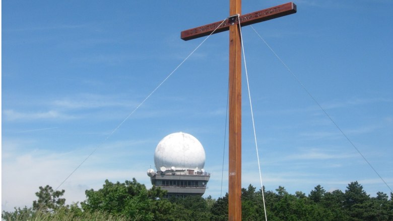 Ein Holzkreuz vor einer Radarkuppel auf dem Buschberg, umgeben von Bäumen und blauem Himmel.