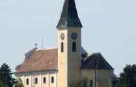 Pfarrkirche Großebersdorf mit spitzem Turm und Uhr, umgeben von Bäumen.