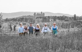 Eine Gruppe von sechs Personen in traditioneller Kleidung steht auf einem Feld mit einer Burg im Hintergrund.