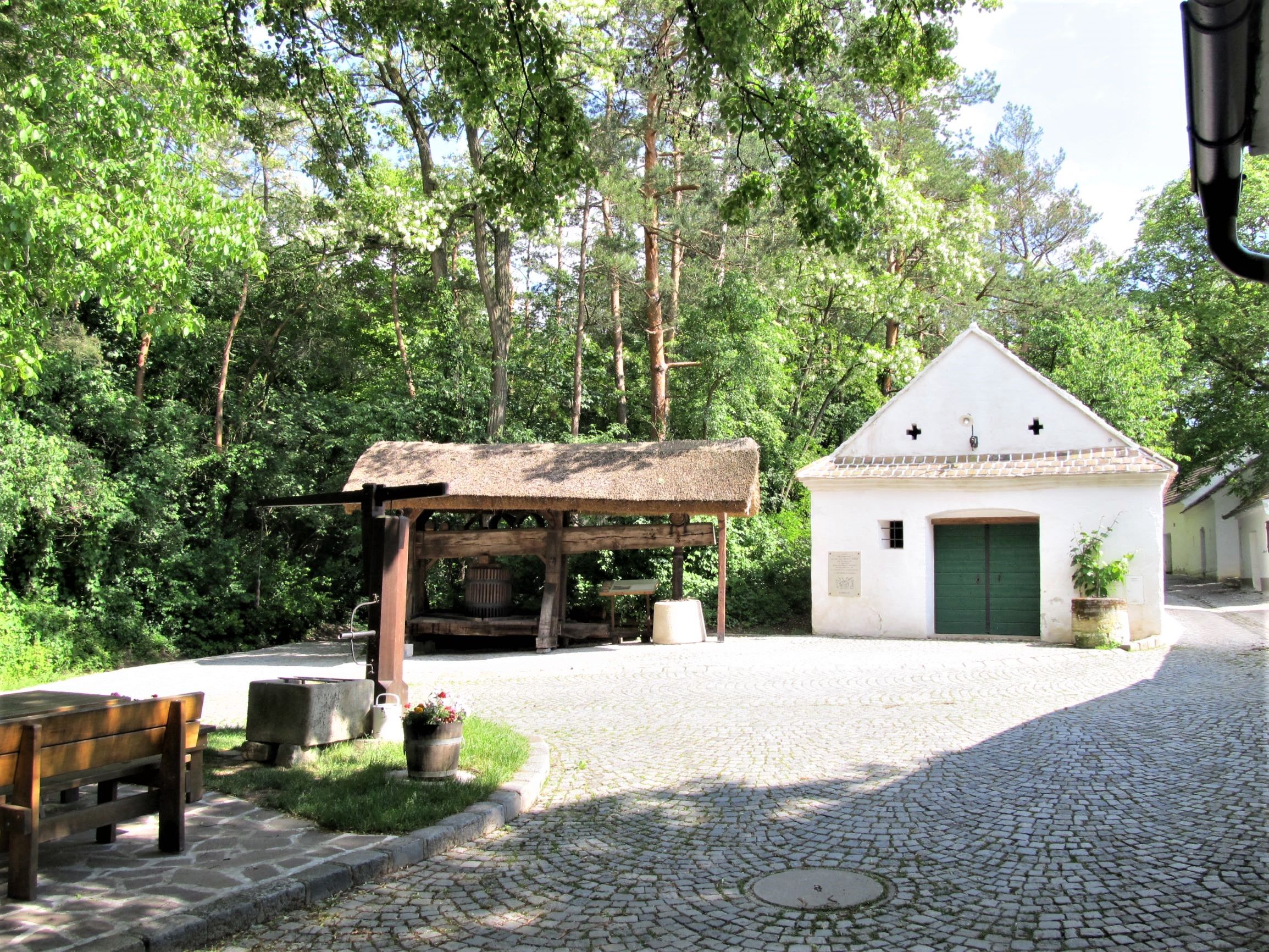 Ein traditioneller Weinkeller mit einer alten Weinpresse und einem kleinen weißen Gebäude im Hintergrund, umgeben von Bäumen.