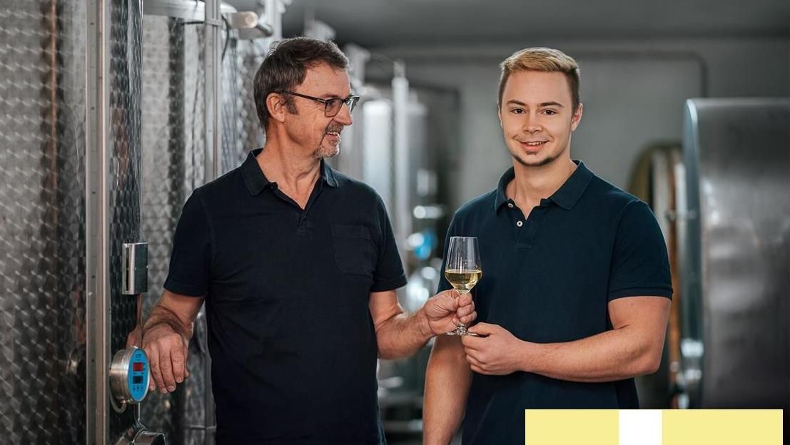 Dvaja muži vo vínnej pivnici, jeden drží pohár bieleho vína, druhý podáva cisternu.