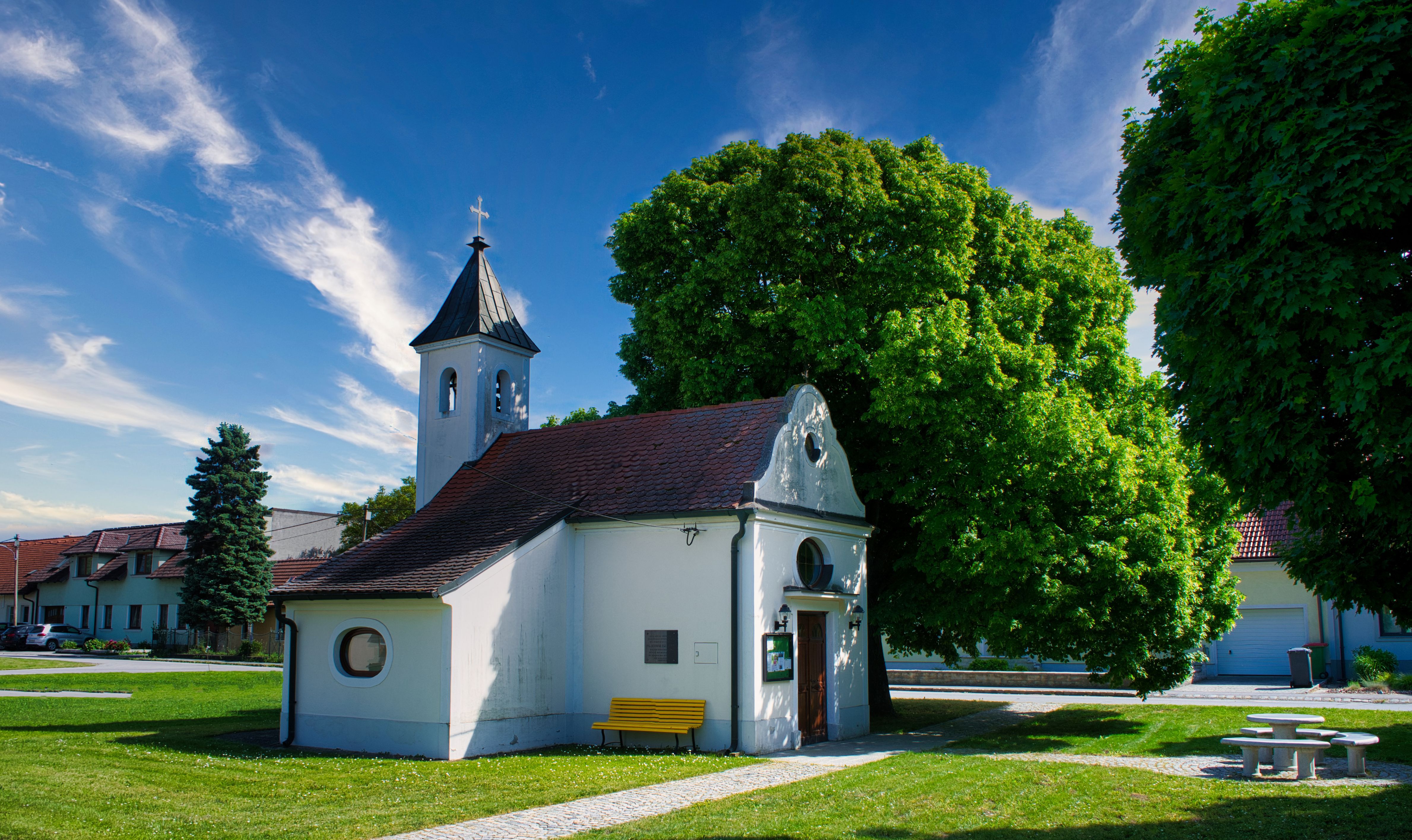 Gemeinde Großhofen: kleine weiße Kapelle mit Glockenturm und Kreuz, umgeben von Bäumen und Grün unter blauem Himmel.