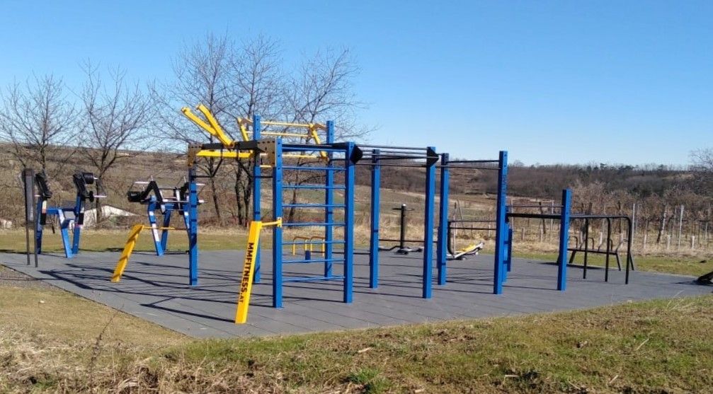 Calisthenics-Park im Freien mit blauen und gelben Geräten, umgeben von einer ländlichen Landschaft.