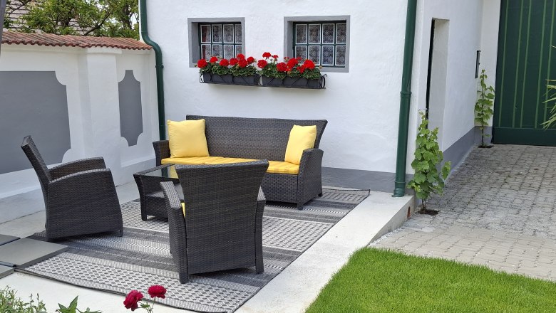 Terrasse mit Rattanmöbeln und gelben Kissen, umgeben von weißen Wänden und roten Blumen.