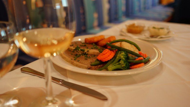 Ein Teller mit Fleisch, Gemüse und Kartoffeln auf einem Tisch, daneben ein Glas Weißwein.