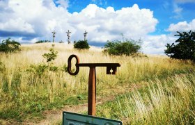 Ein Fotopunkt-Schild vor einer Wiese mit drei Kreuzen im Hintergrund unter blauem Himmel mit Wolken.