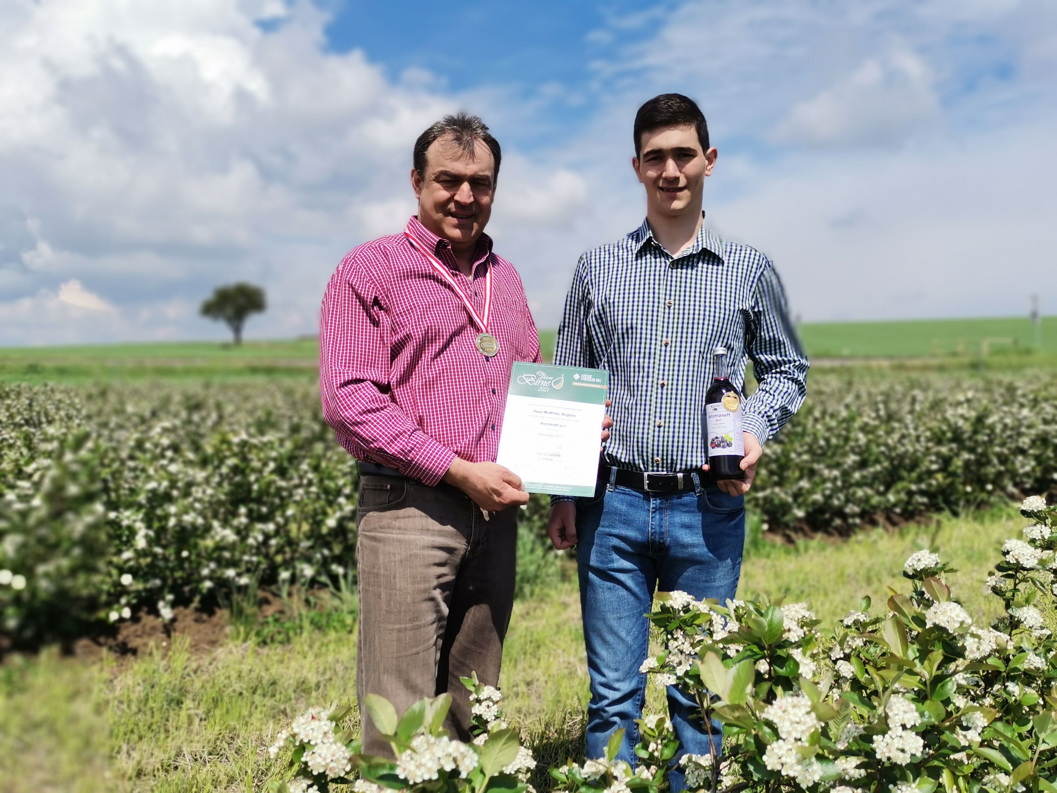 Zwei Männer stehen in einem blühenden Feld, einer hält eine Urkunde und eine Medaille, der andere eine Flasche.