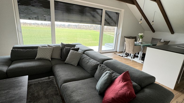 Wohnzimmer mit grauem Sofa, rotem Kissen, Esstisch und Panoramafenster mit Blick auf gr&uuml;ne Wiese.