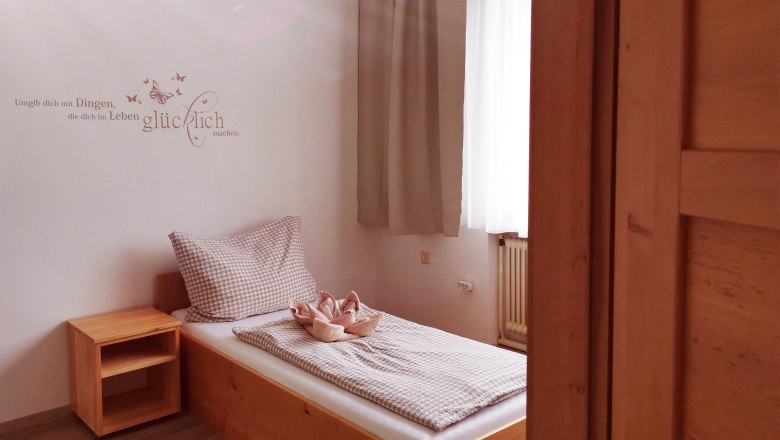 Ein einfaches Gästezimmer mit einem Einzelbett, Holzregal und einem Zitat an der Wand.