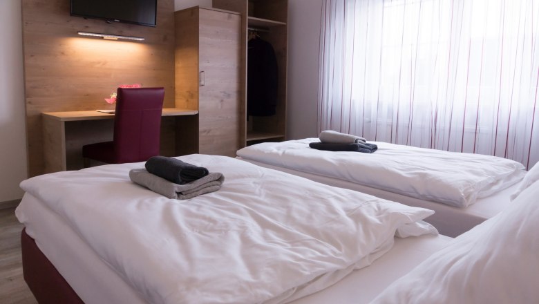 Zweibettzimmer mit zwei Betten, Schreibtisch, Stuhl und Kleiderschrank. Handt&uuml;cher auf den Betten, Fenster mit Vorh&auml;ngen im Hintergrund.