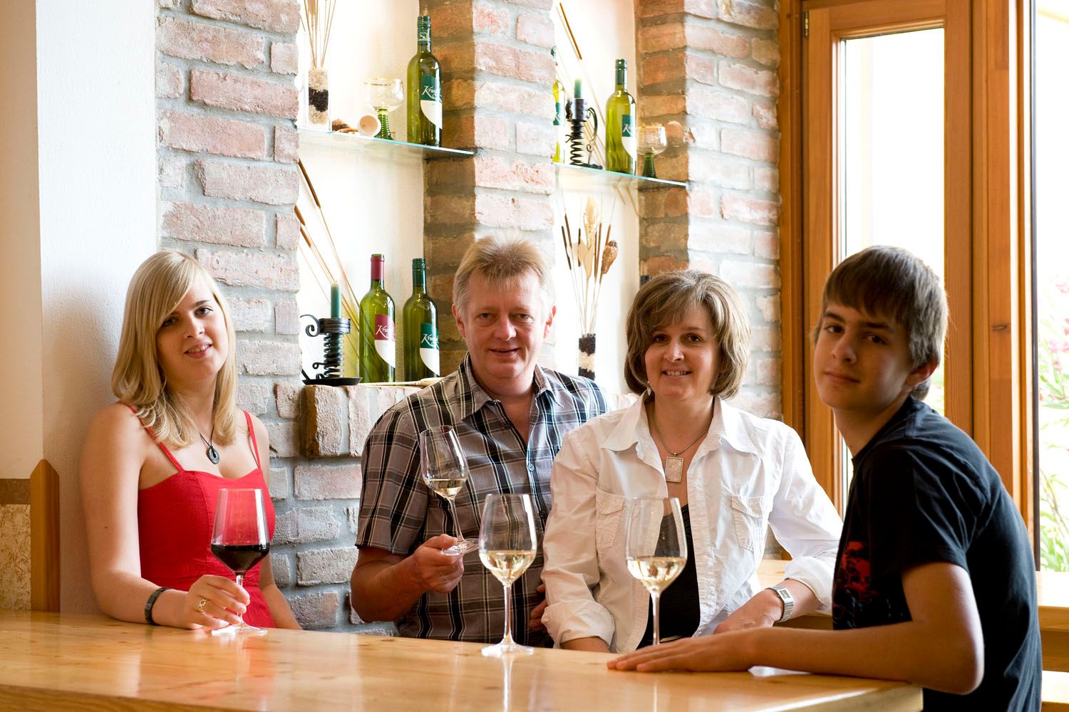 Vier Personen sitzen an einem Tisch mit Weingläsern, im Hintergrund Weinflaschen auf Regalen.