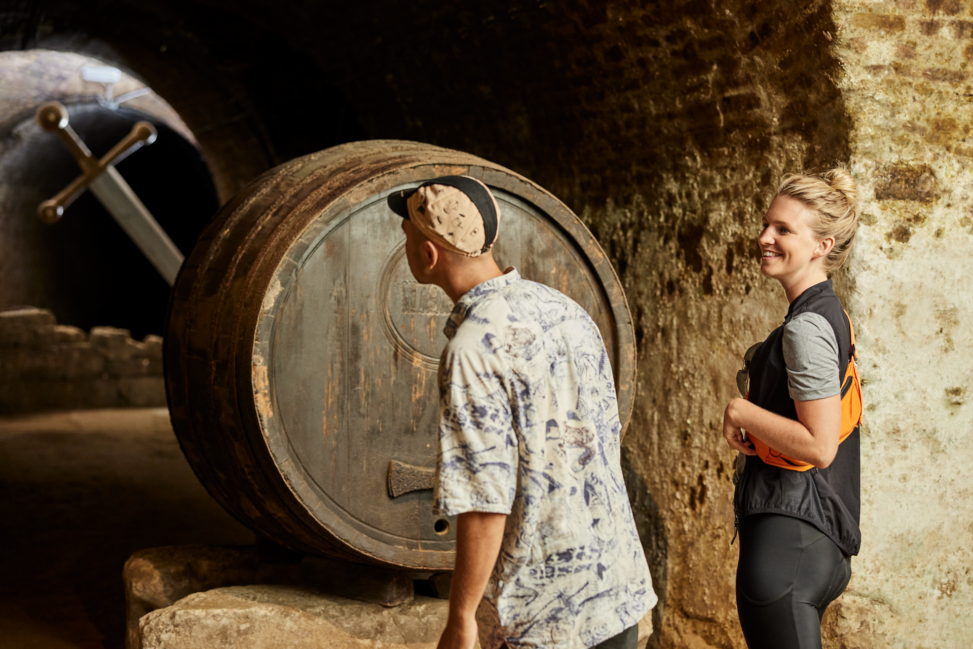In einem historischen Kellergewölbe, umgeben von großen Holzfässern, genießen Besucher die Atmosphäre des Weinviertels. Ein Lächeln und angeregte Gespräche schaffen eine einladende Stimmung, während die rustikale Einrichtung die Tradition der Weinproduktion widerspiegelt.
