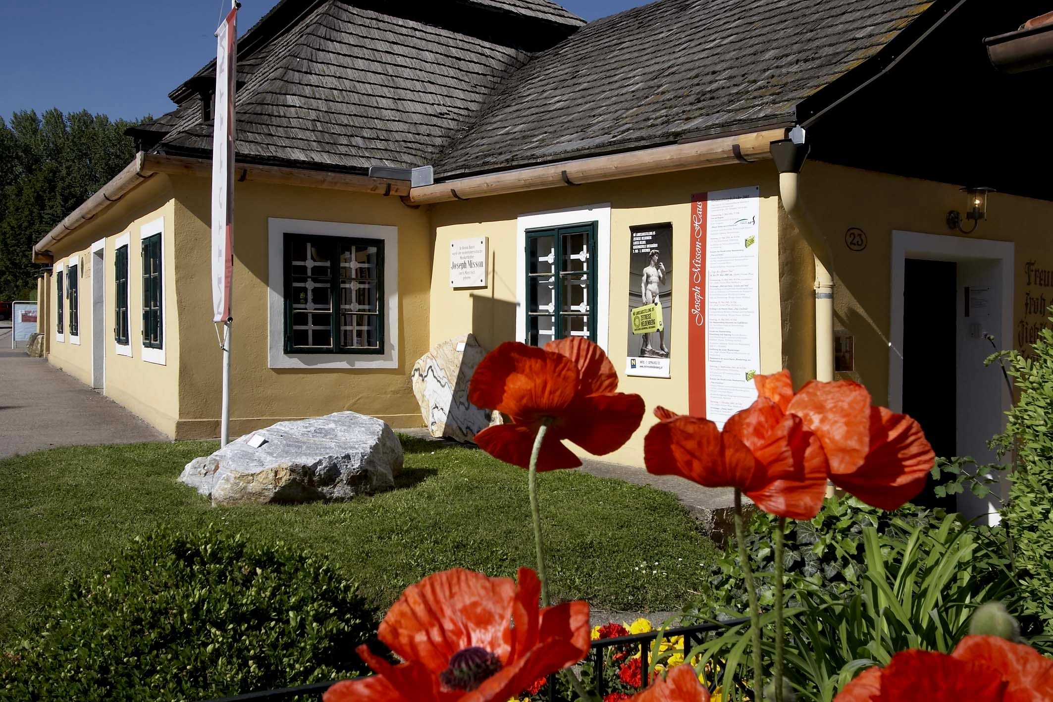 Gelbes Haus mit Schindeldach und Mohnblumen im Vordergrund.