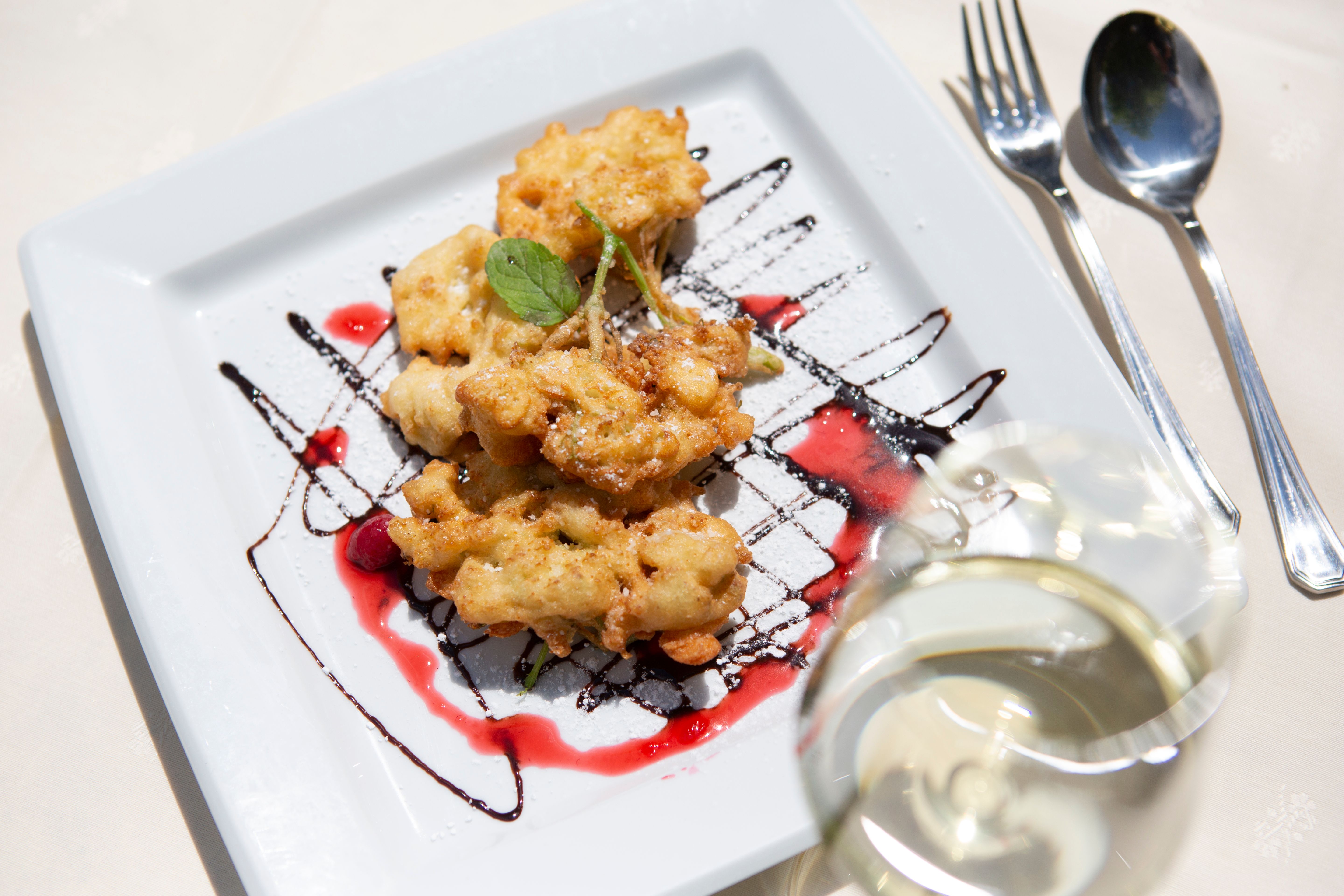 Ein Teller mit frittierten Blüten, garniert mit Schokoladen- und Fruchtsauce, auf einem weißen Teller. Ein Glas Weißwein steht daneben.