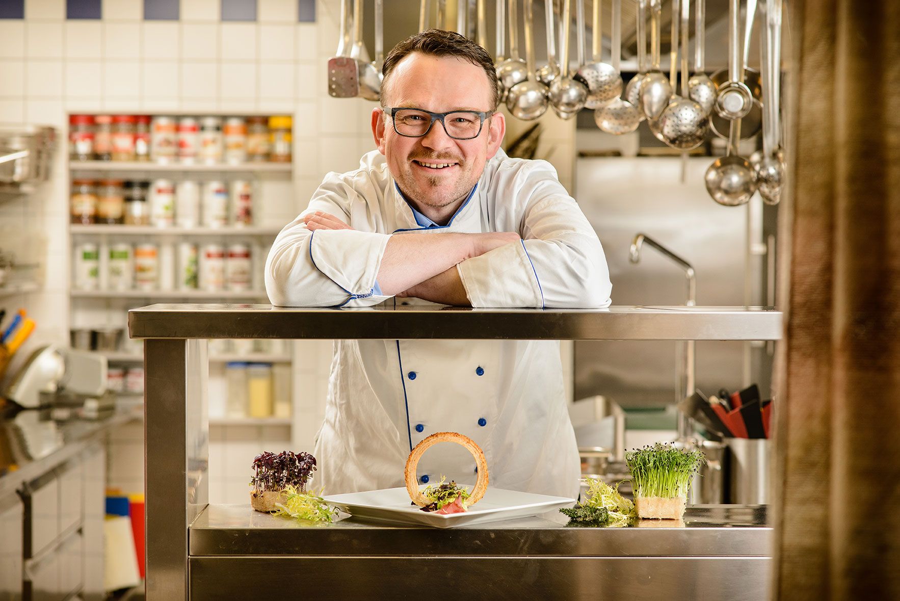 A chef in a white uniform leans on a stainless steel counter in a kitchen. In front of him is a plate of decorative food.