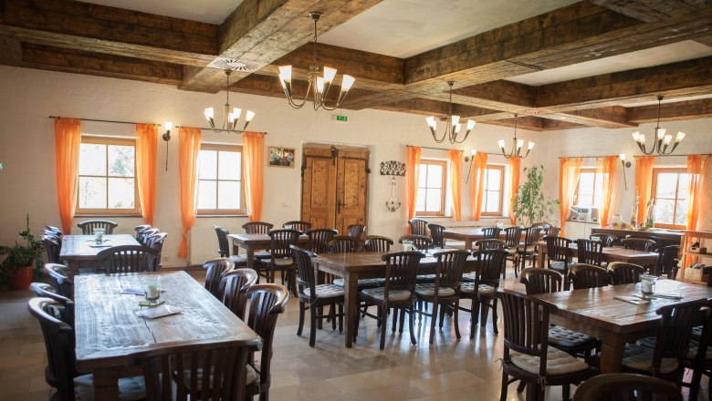 A rustic dining room with wooden tables and chairs, orange curtains and a wooden beamed ceiling.