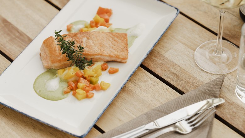 Ein Teller mit gebratenem Lachs auf Gemüse und grüner Sauce, dekoriert mit einem Kräuterzweig, auf einem Holztisch mit Besteck und Weinglas.